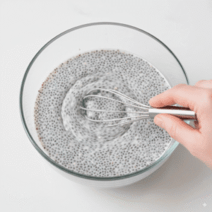 A hand using a whisk to stir a partially gelled chia seed pudding in a glass bowl, the key step to prevent clumps.