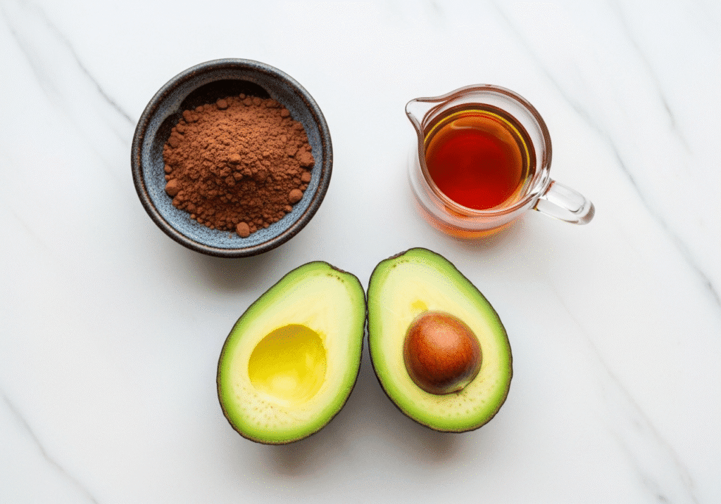 An overhead view of the simple ingredients for a chocolate avocado mousse recipe: halved avocados, cocoa powder, and maple syrup.