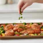 A hand sprinkling fresh parsley over a finished sheet pan of lemon herb chicken and roasted vegetables.